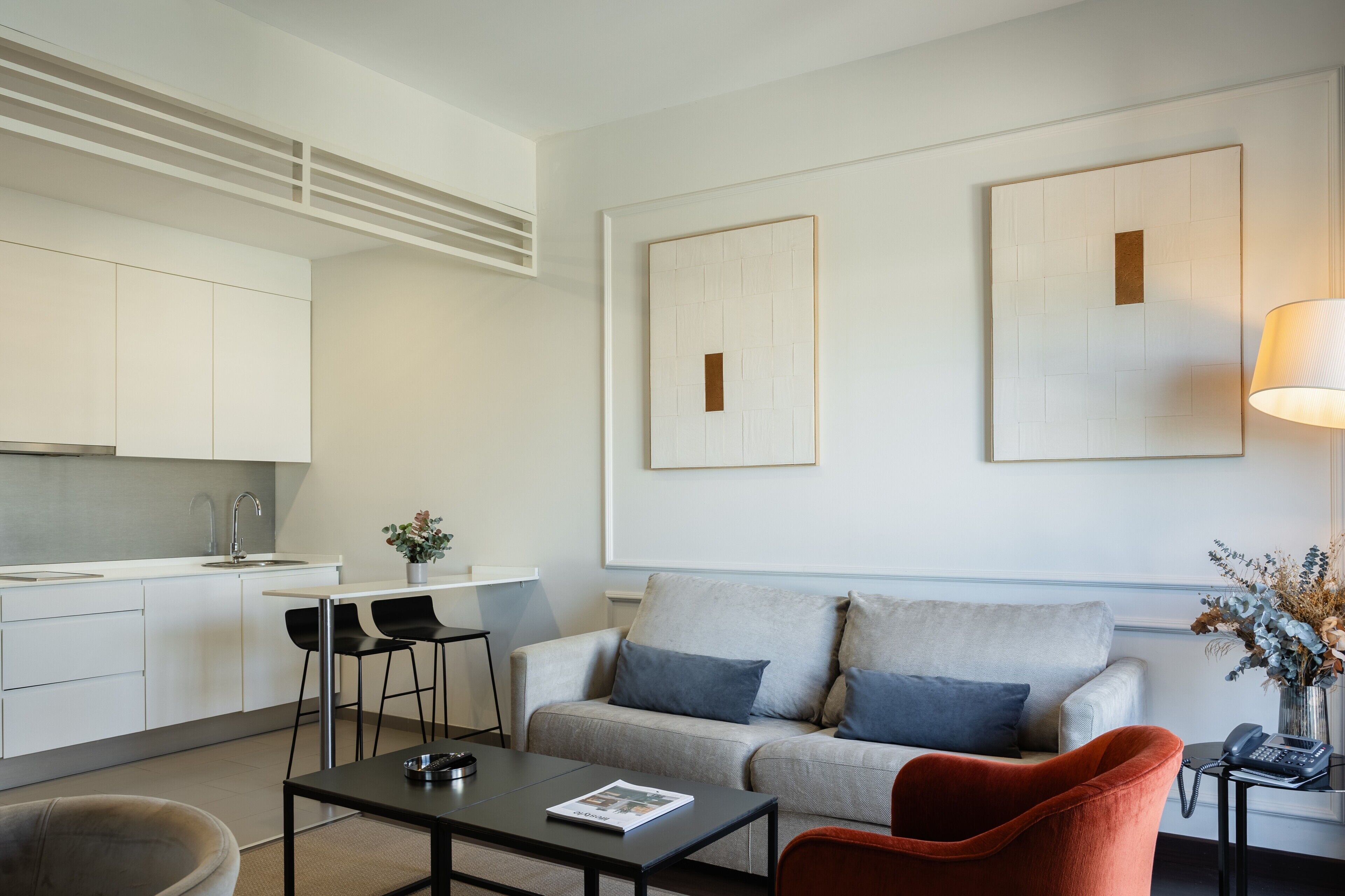 A modern living room with a sofa, coffee table, and chairs. There is a kitchen area with a sink and cabinets.
