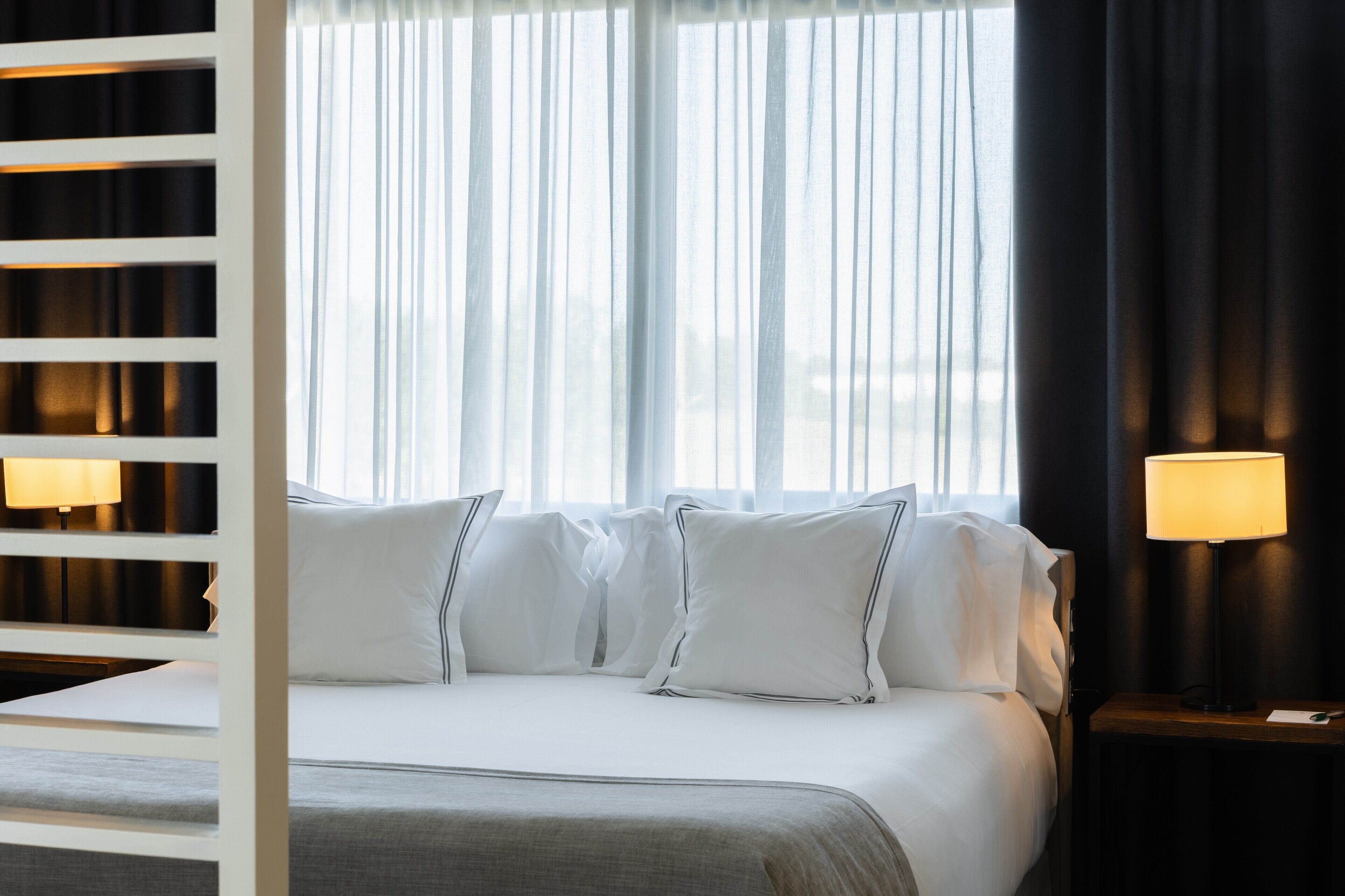 Una habitación de hotel con una cama, dos lámparas de noche y una ventana con cortinas de gasa.
