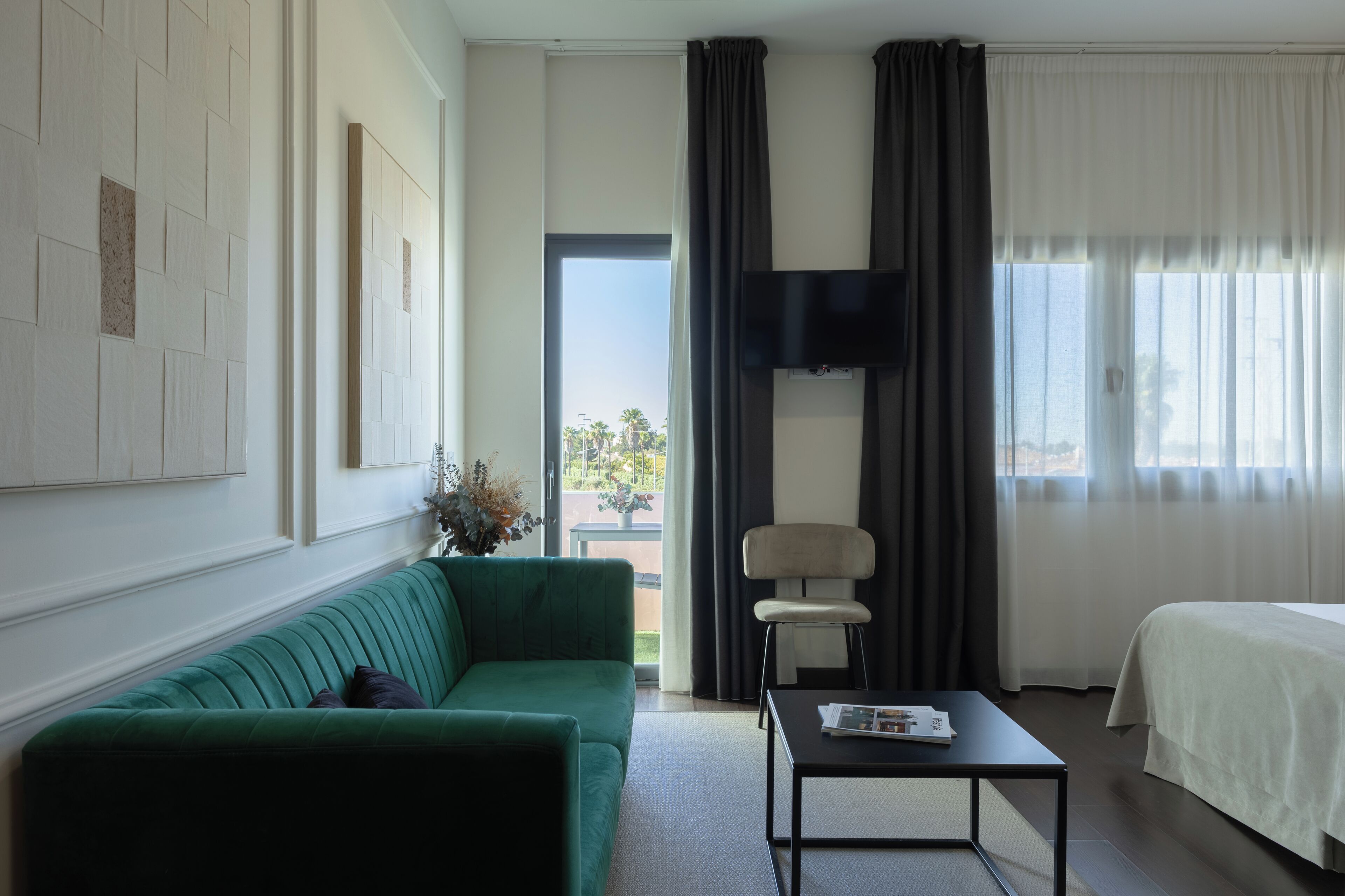 Una habitación de hotel con un sofá verde, una cama, un televisor de pantalla plana montado en la pared, una mesita con una revista y una ventana con cortinas.