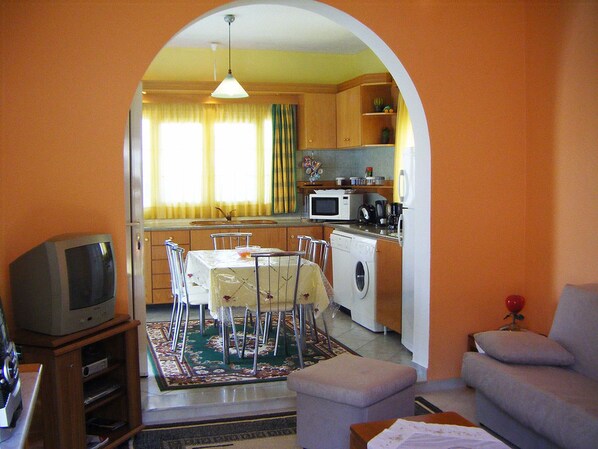 A living room with a television, a sofa, a dining table with chairs, and a kitchen area with appliances.
