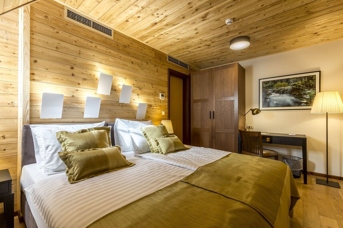A wooden-paneled bedroom with a bed, bedside lamps, a desk, and a framed picture on the wall.
