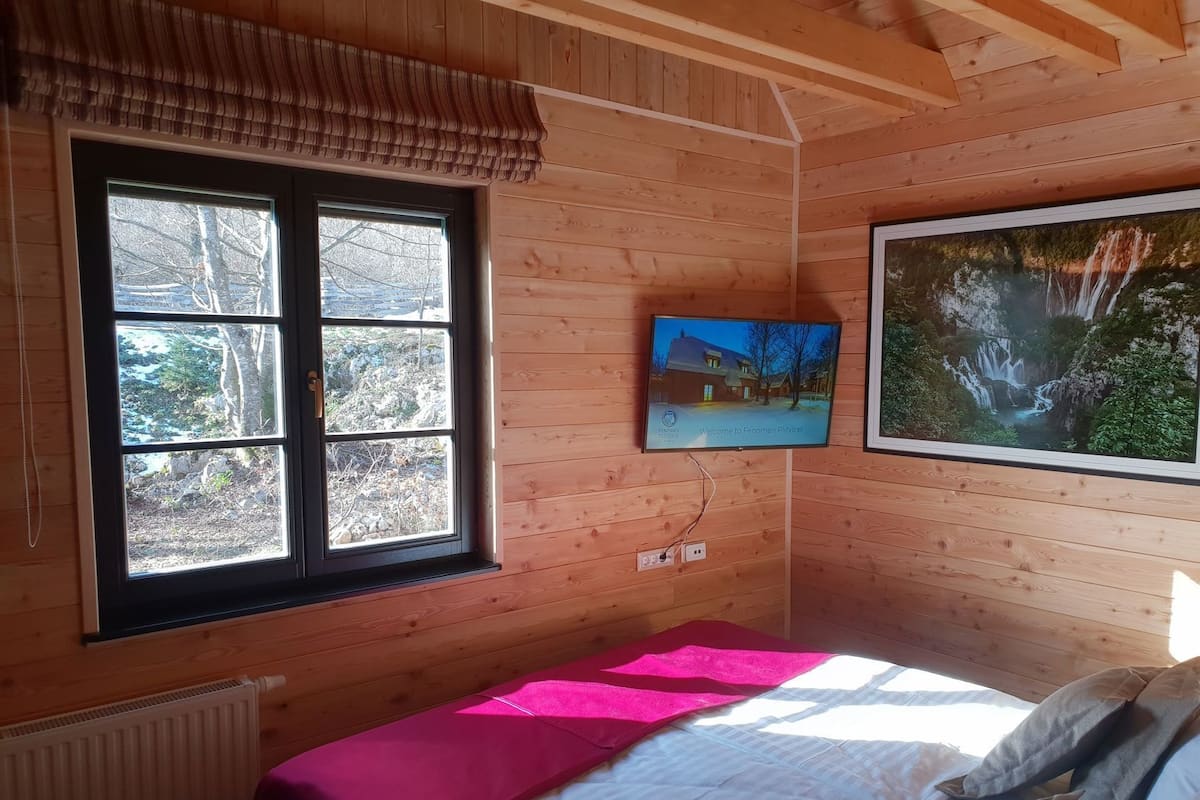 Ein Zimmer einer Holzhütte mit einem Bett, einem Fenster mit Blick auf Bäume und einem Fernseher, der ein Haus zeigt.