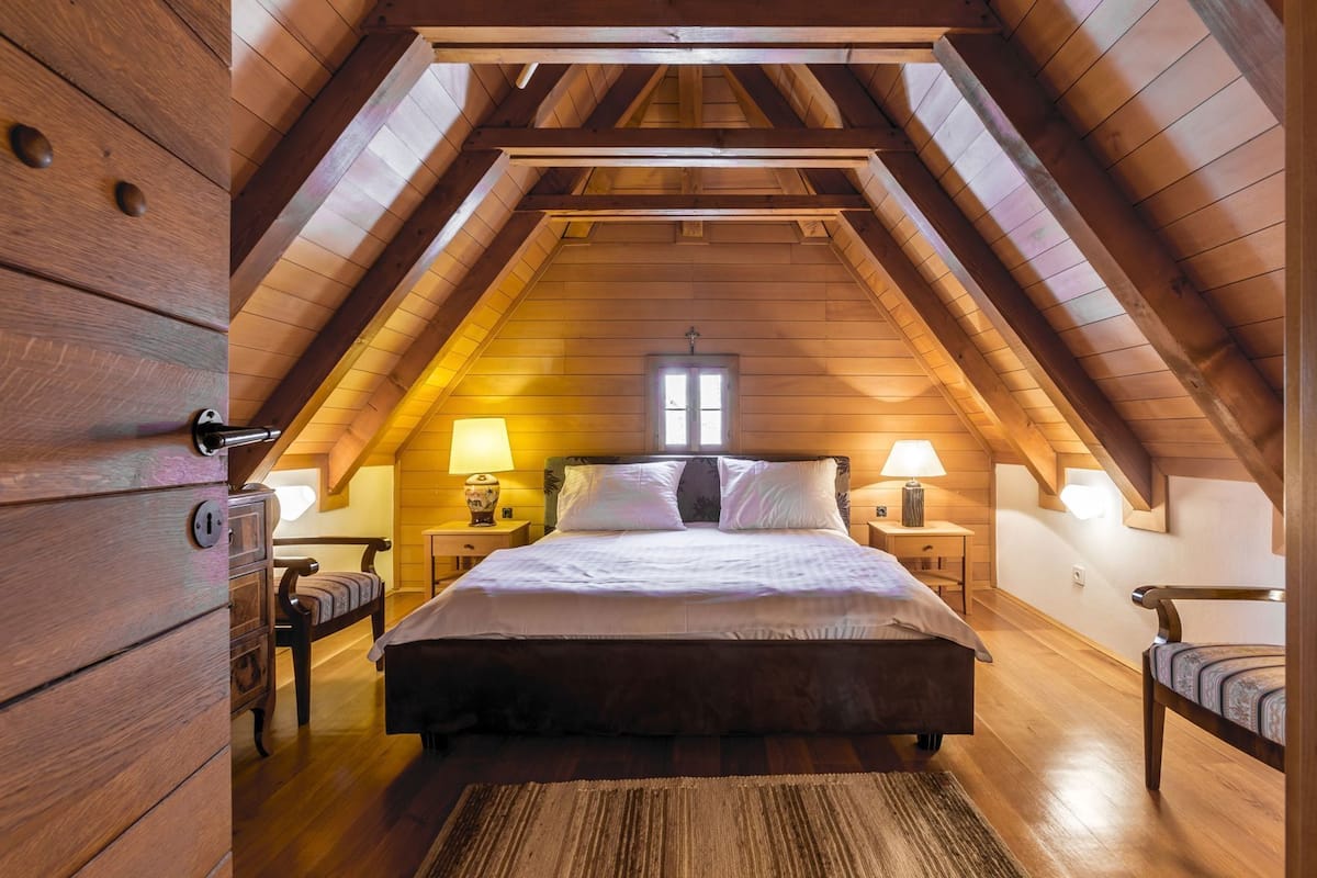 Ein Schlafzimmer mit schrägen Wänden, Holzwänden, einem Bett mit Kissen, zwei Nachttischen mit Lampen und einem Fenster.