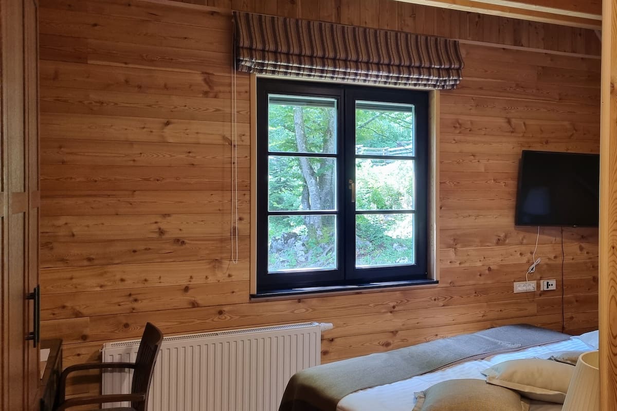 Ein Zimmer in einer Holzhütte mit Bett, Stuhl, Fenster mit Blick auf Bäume und einem an der Wand befestigten Fernseher.
