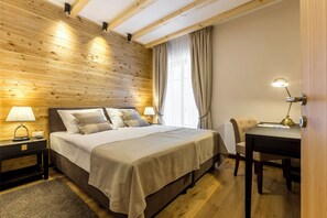 A bedroom with a wooden ceiling, a bed with white bedding, a nightstand with a lamp, a chair, and a table with a lamp.
