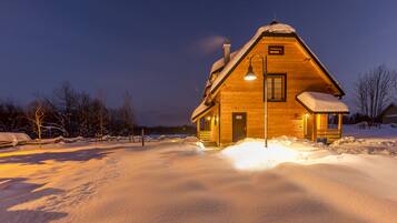 A wooden cabin with a sloped roof and a lit entrance, surrounded by snow.