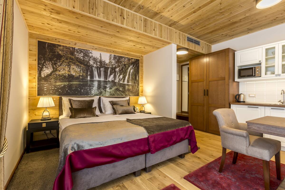 Ein Schlafzimmer mit einem Bett, Nachttischlampen, einem großen Gemälde eines Wasserfalls, einer Holzzielung und einer Küchenzeile mit einer Mikrowelle.