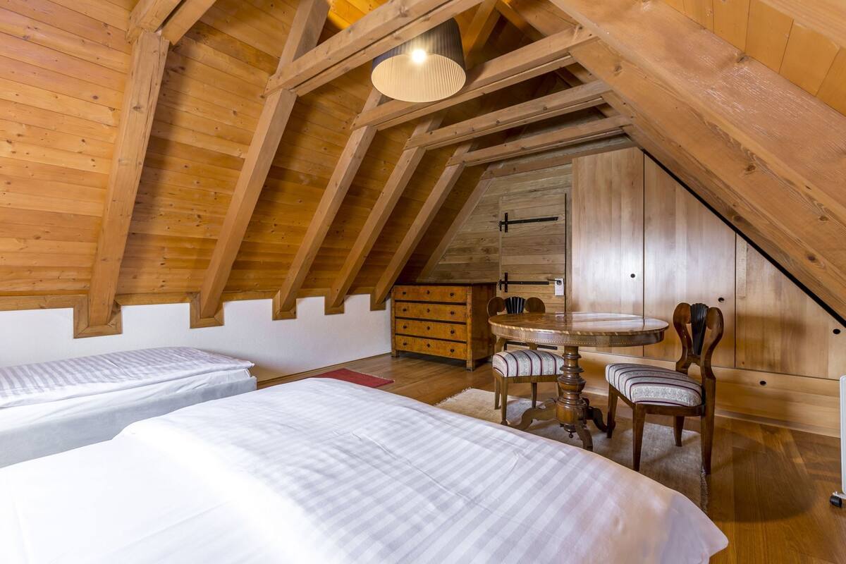 Ein Schlafzimmer im Loft-Stil mit zwei Betten, einer Holzkommode, einem runden Tisch mit Stühlen und einer schrägen Decke.