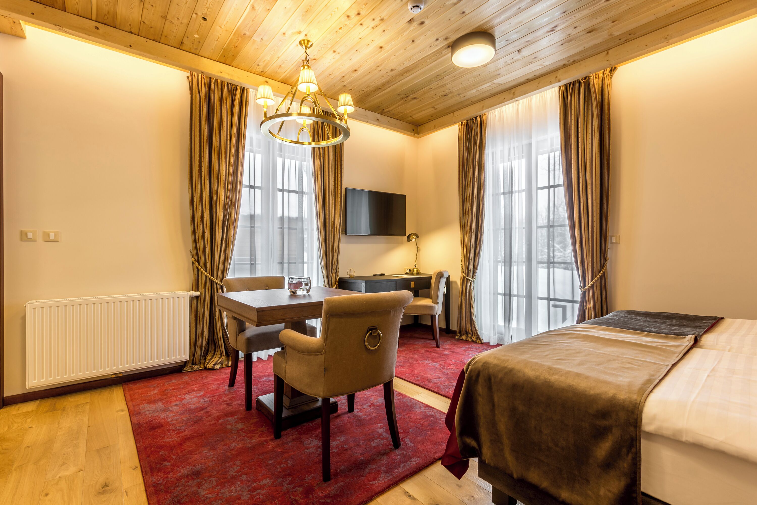 A hotel room with a wooden ceiling, a red carpet, a bed, a desk with a chair, a TV mounted on the wall, and a window with curtains.