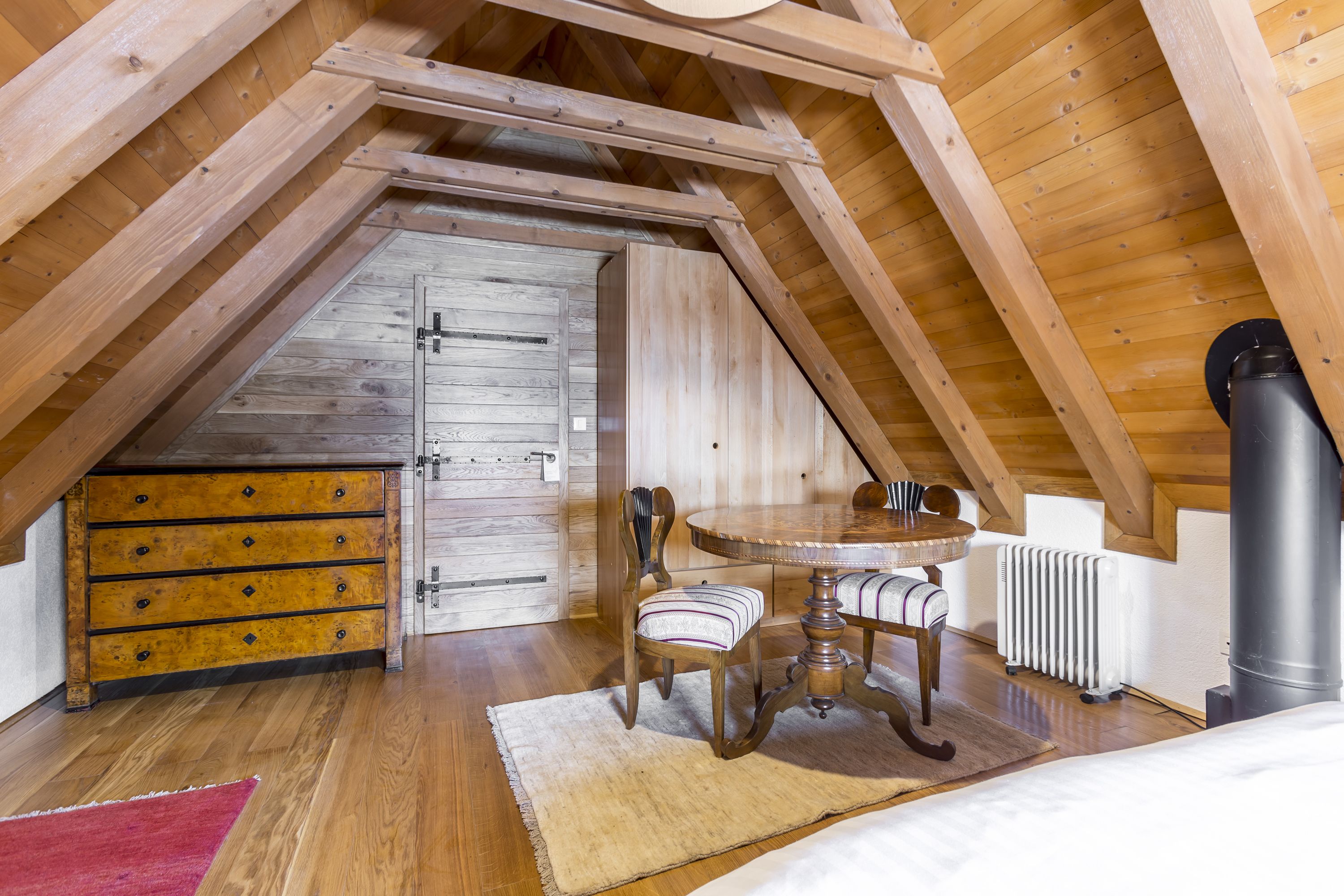 Une chambre mansardée chaleureuse avec des poutres apparentes, une commode en bois, une table de salle à manger ronde, une chaise et un radiateur.