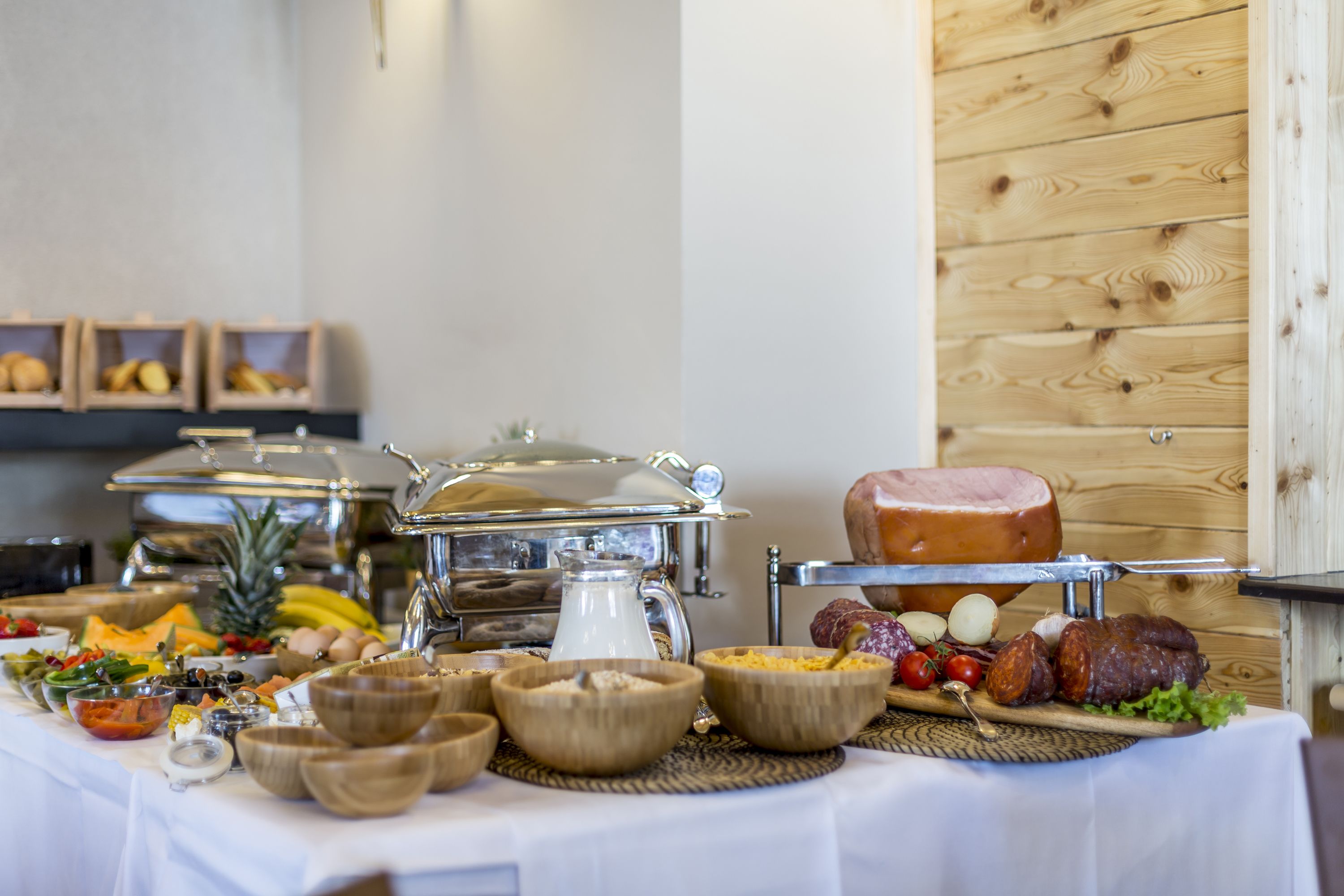 Una mesa de buffet con diversos alimentos, como frutas, verduras y carnes, en una sala con paredes de madera y un mantel blanco.