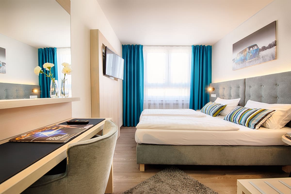 A hotel room with a bed, a desk with a chair, a TV, and a window with curtains.