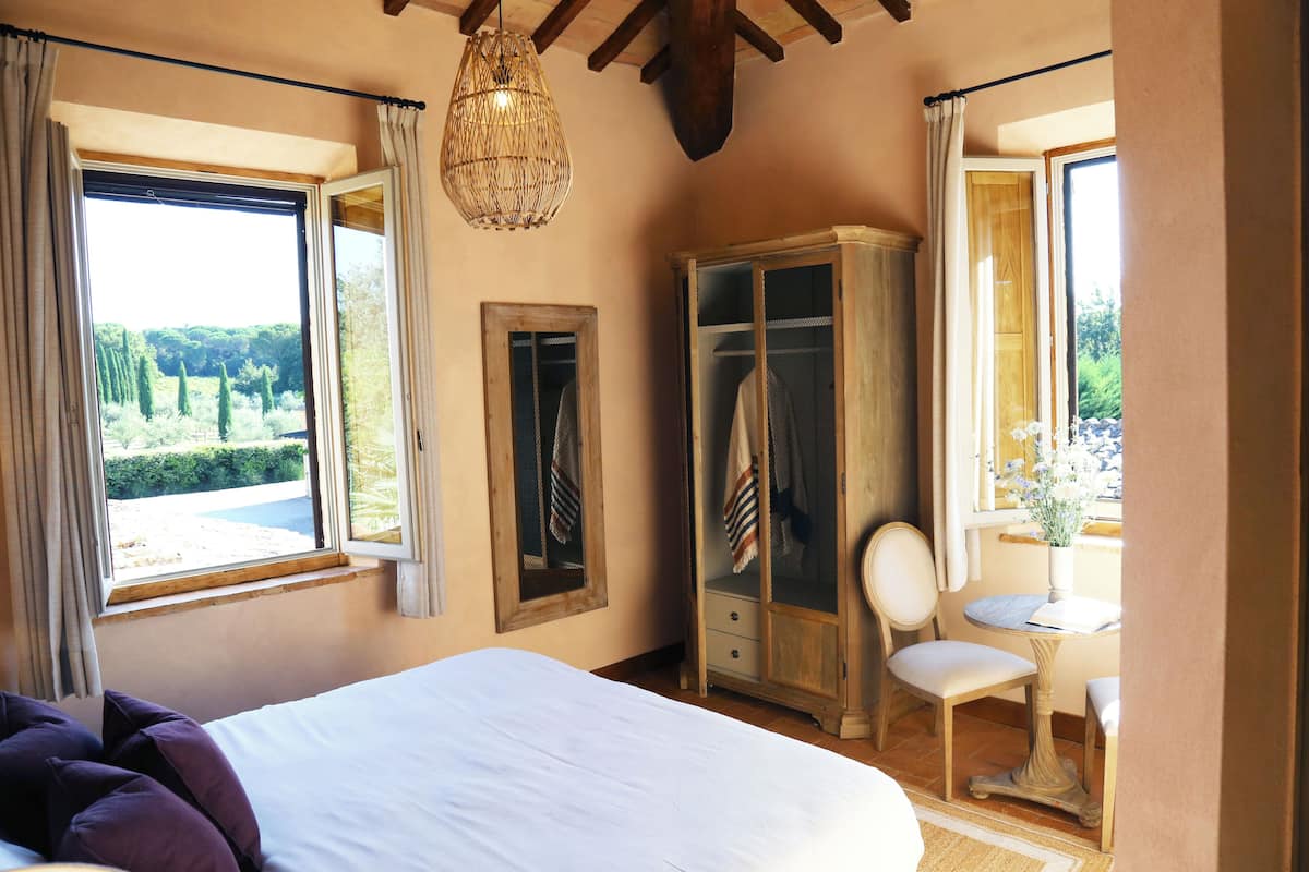 Een slaapkamer met een groot bed, een kledingkast, een stoel en een tafel bij een raam met uitzicht op groen.
