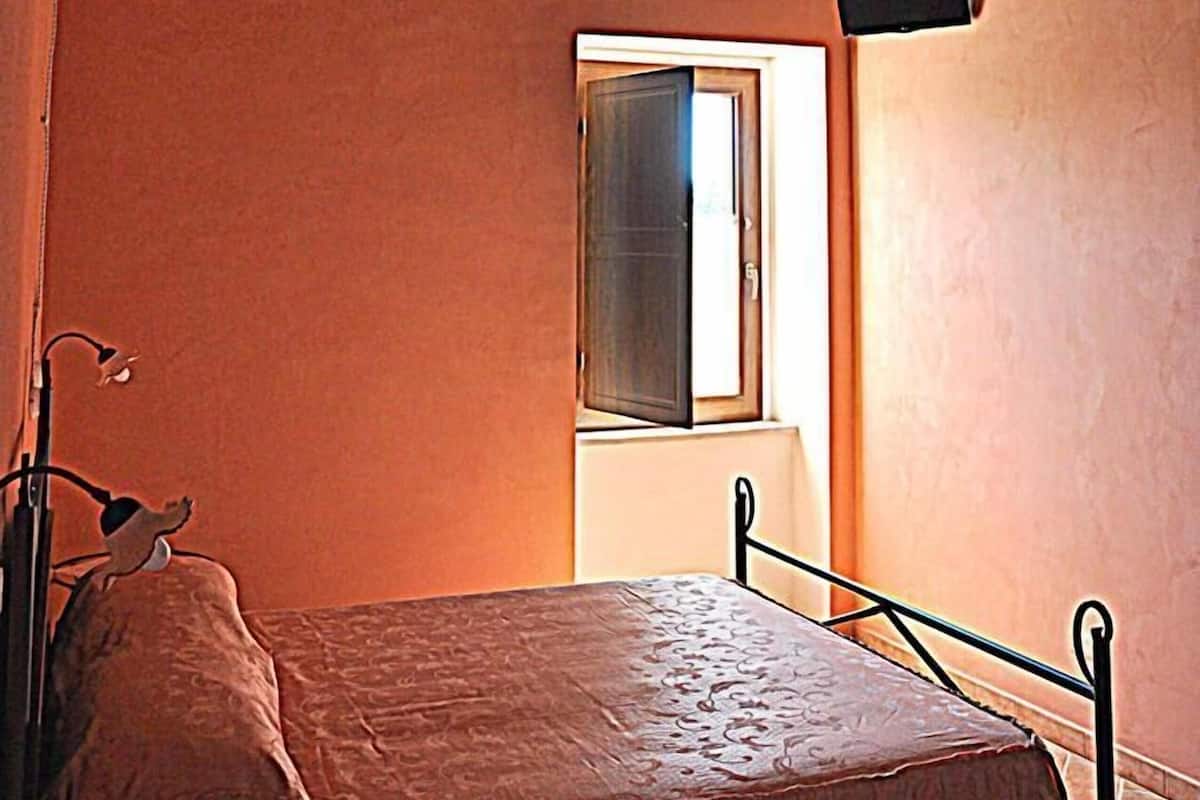 A bedroom with a single bed, wooden ceiling beams, a small window, and a wall-mounted speaker.