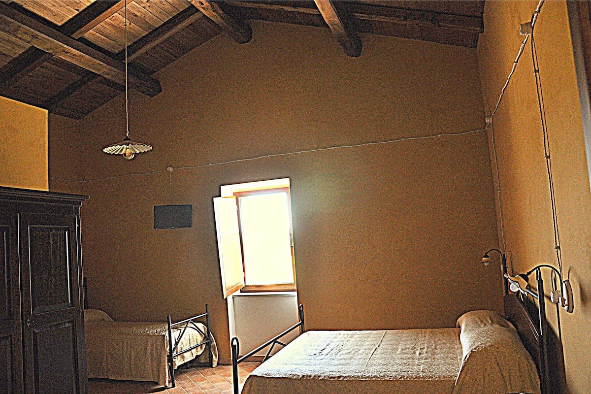A room with a bed, a chair, a wooden ceiling, a window, and a wall-mounted light fixture.