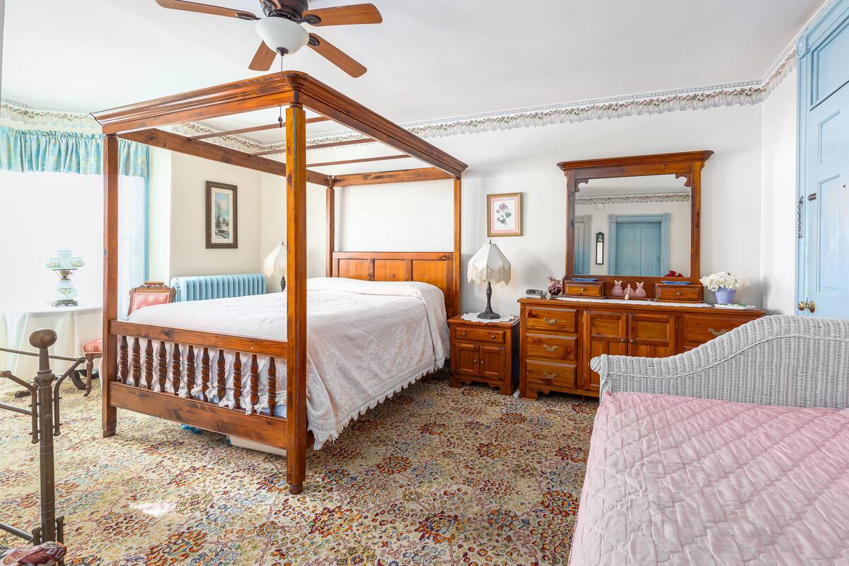 A four-poster bed with a wooden frame, a nightstand with a lamp, a dresser with a mirror, and a patterned carpet.