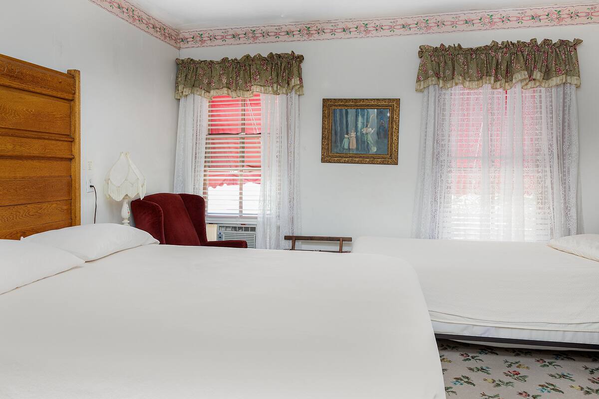 A bedroom with two beds, a wooden wardrobe, a red chair, and a painting on the wall.