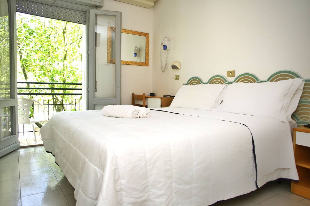 A hotel room with a bed, a desk, a chair, a mirror, and a view of greenery outside.