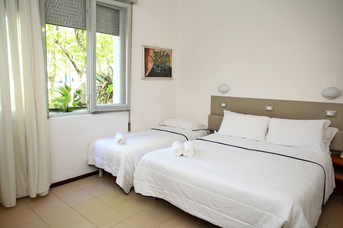A hotel room with two beds, a window with curtains, a painting on the wall, and a view of greenery outside.