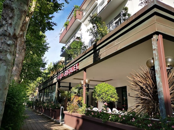 A hotel named "HOTEL RISTALEX" with a modern facade, outdoor plants, and a clear sky.