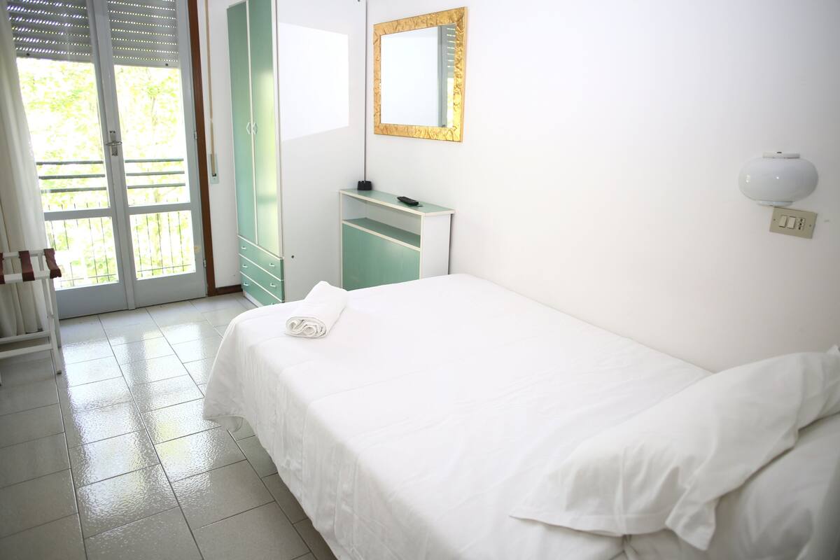 A hotel room with a bed, a desk, a mirror, and a door leading to an outdoor area.