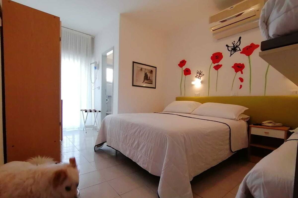 A hotel room with a bed, a bedside table, a chair, and a small dog.