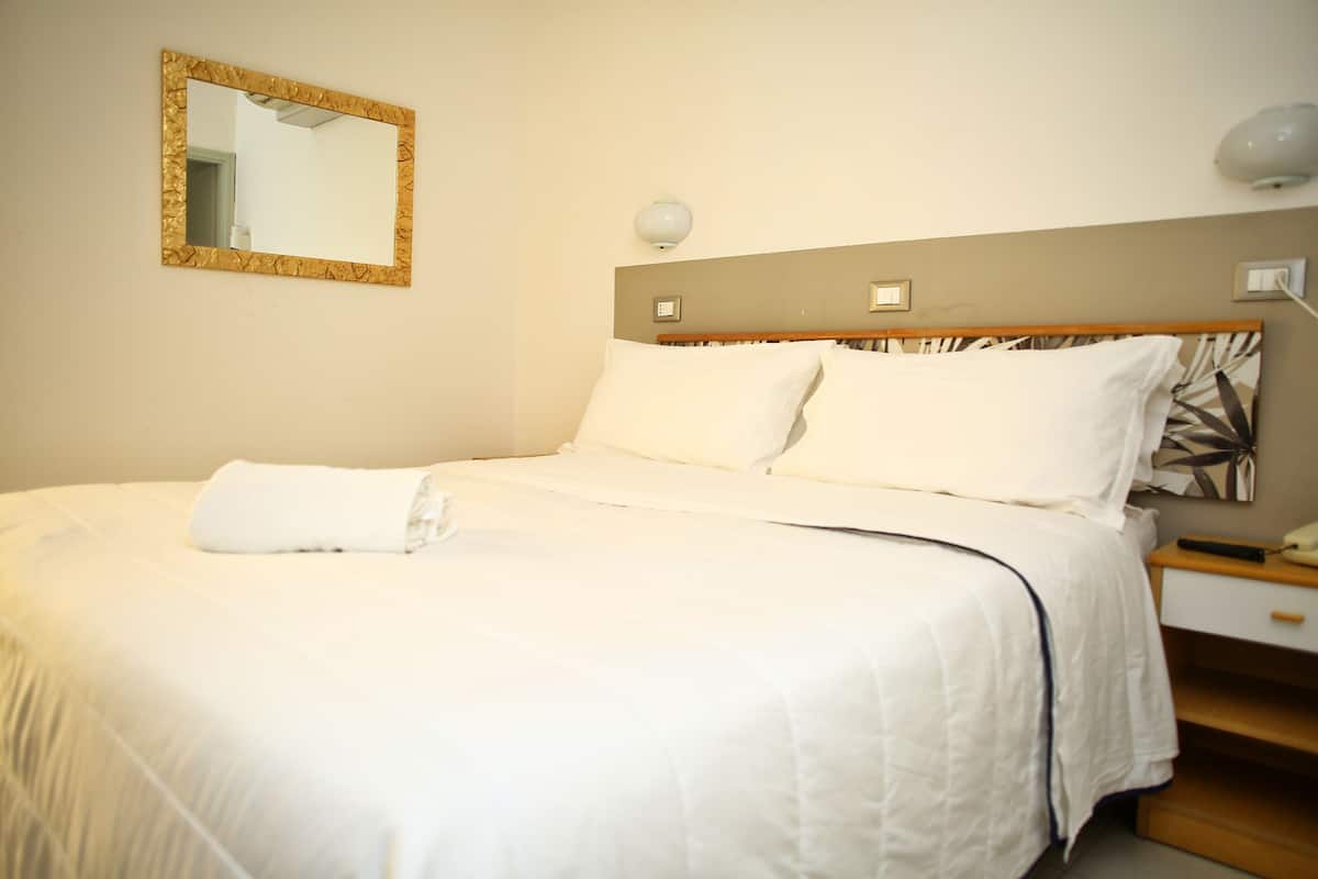 A neatly made bed with white linens, a bedside table with a lamp, and a wall-mounted mirror.