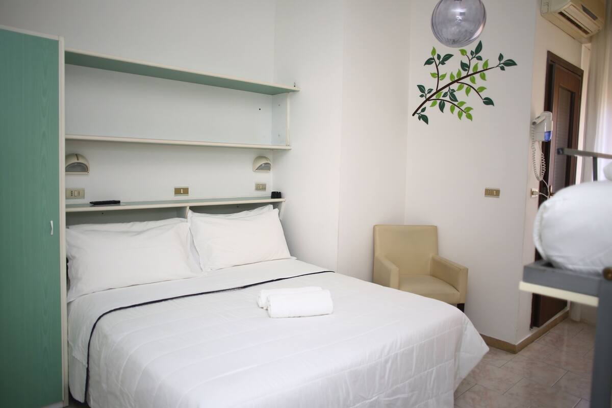 A hotel room with a bed, a chair, a shelf, and a wall-mounted air conditioning unit.