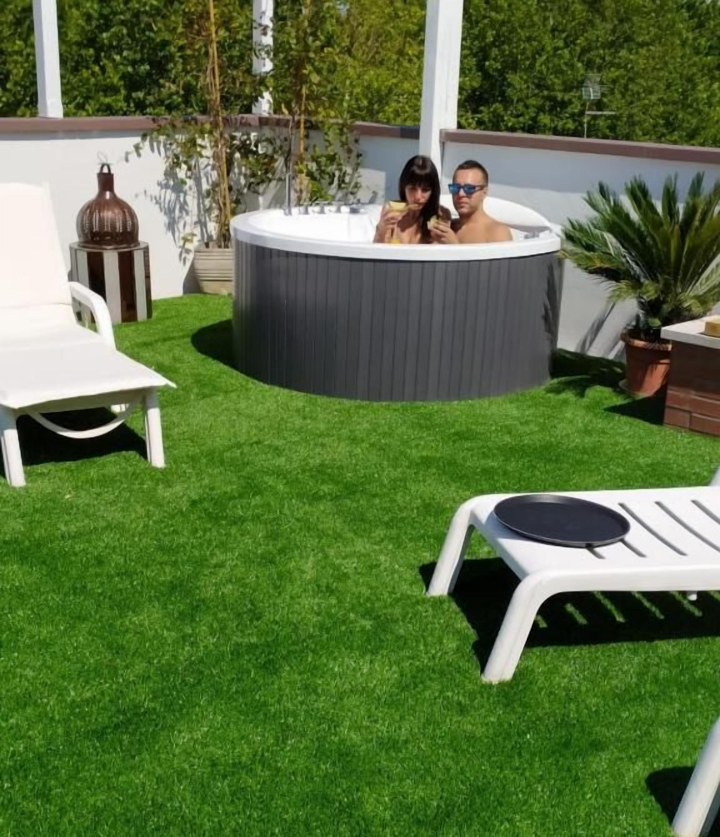 A hot tub surrounded by artificial grass, white lounge chairs, and potted plants.