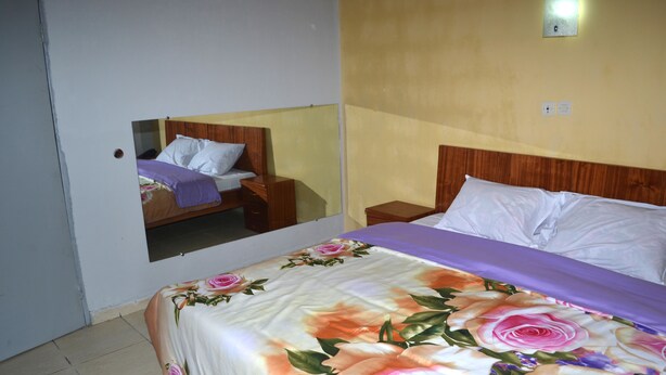 A hotel room with a bed, a nightstand, a mirror, and a wall-mounted light fixture. - Hôtel Ma Résidence (Abidjan)