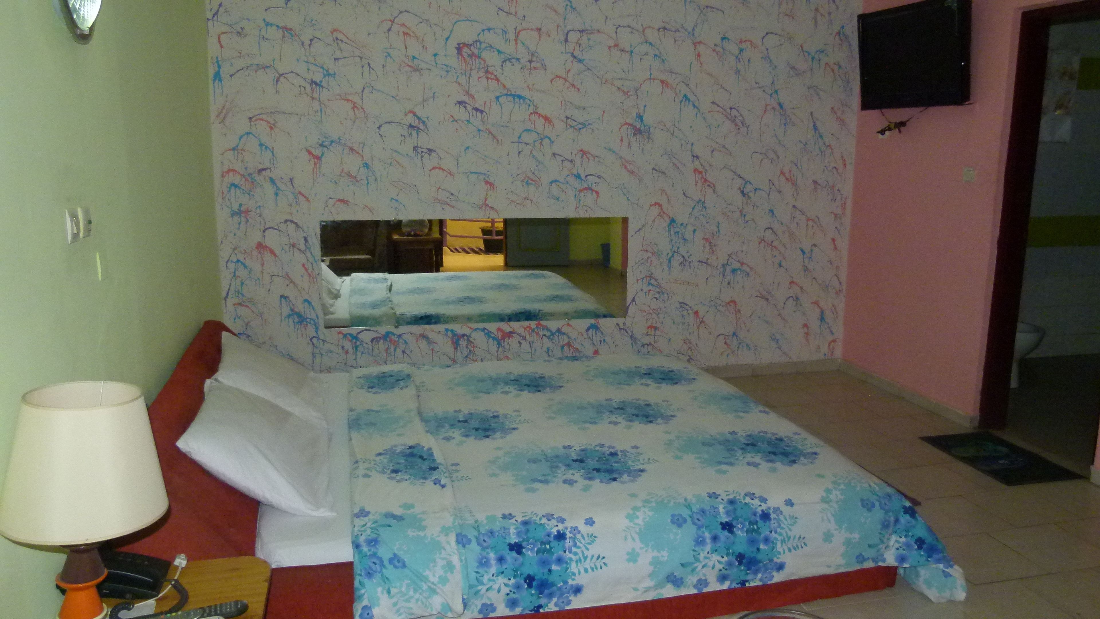 A bedroom with a bed, a bedside table with a lamp, a mirror, and a wall with a pattern.