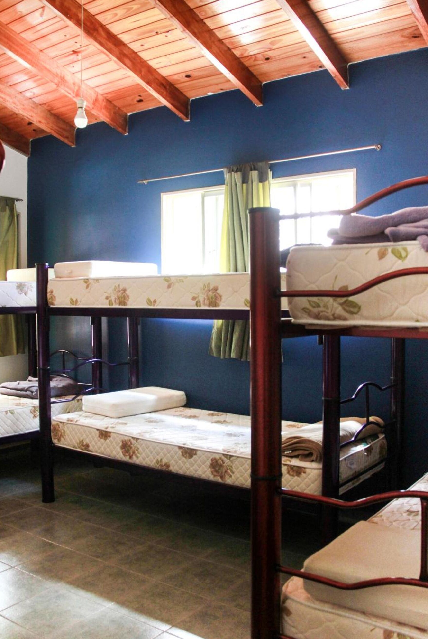 Habitación con literas, vigas de madera, ventana con cortinas y suelo de baldosas.