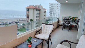 Ein Balkon mit Korbmöbeln, einem kleinen Tisch mit einer Pflanze und Blick auf Strand und Gebäude.