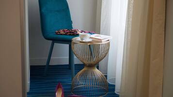 A hotel room with a teal chair, a small round table with a gold wire base, and a pair of purple shoes on the floor.