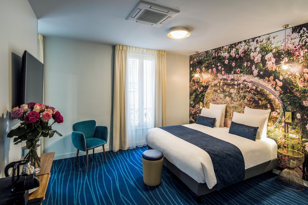 A hotel room with a large bed, a desk with a chair, a vase of flowers, and a wall mural.