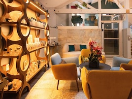 A modern interior with a wooden shelving unit displaying various bottles, a seating area with yellow chairs, and a stone wall.