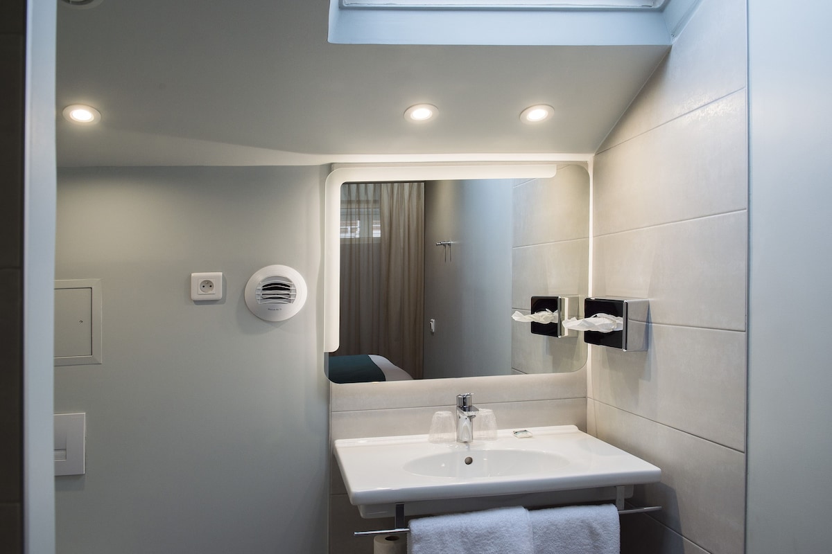 A modern bathroom with a white sink, mirror, and towel rack. There are ceiling lights and a window above the mirror.