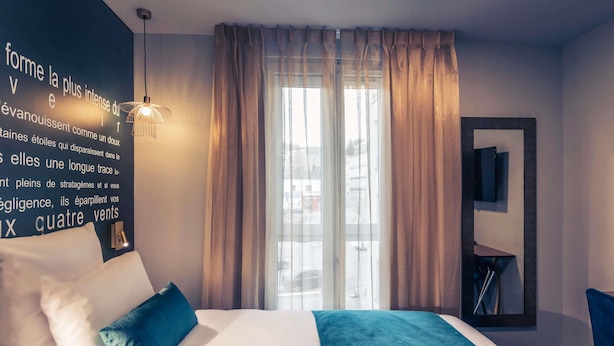 Hotellihuone, jossa on suuri sänky, ikkunasta avautuva näkymä kaupunkiin ja seinä, jossa on ranskankielinen sitaatti.
