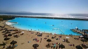 Ein großer Swimmingpool neben einem Sandstrand mit Sonnenschirmen und Liegestühlen.