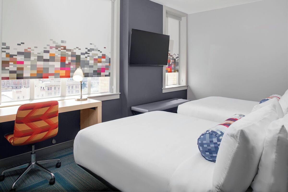 Habitación de hotel con cama, escritorio y silla, televisor y ventana con vista a edificios.