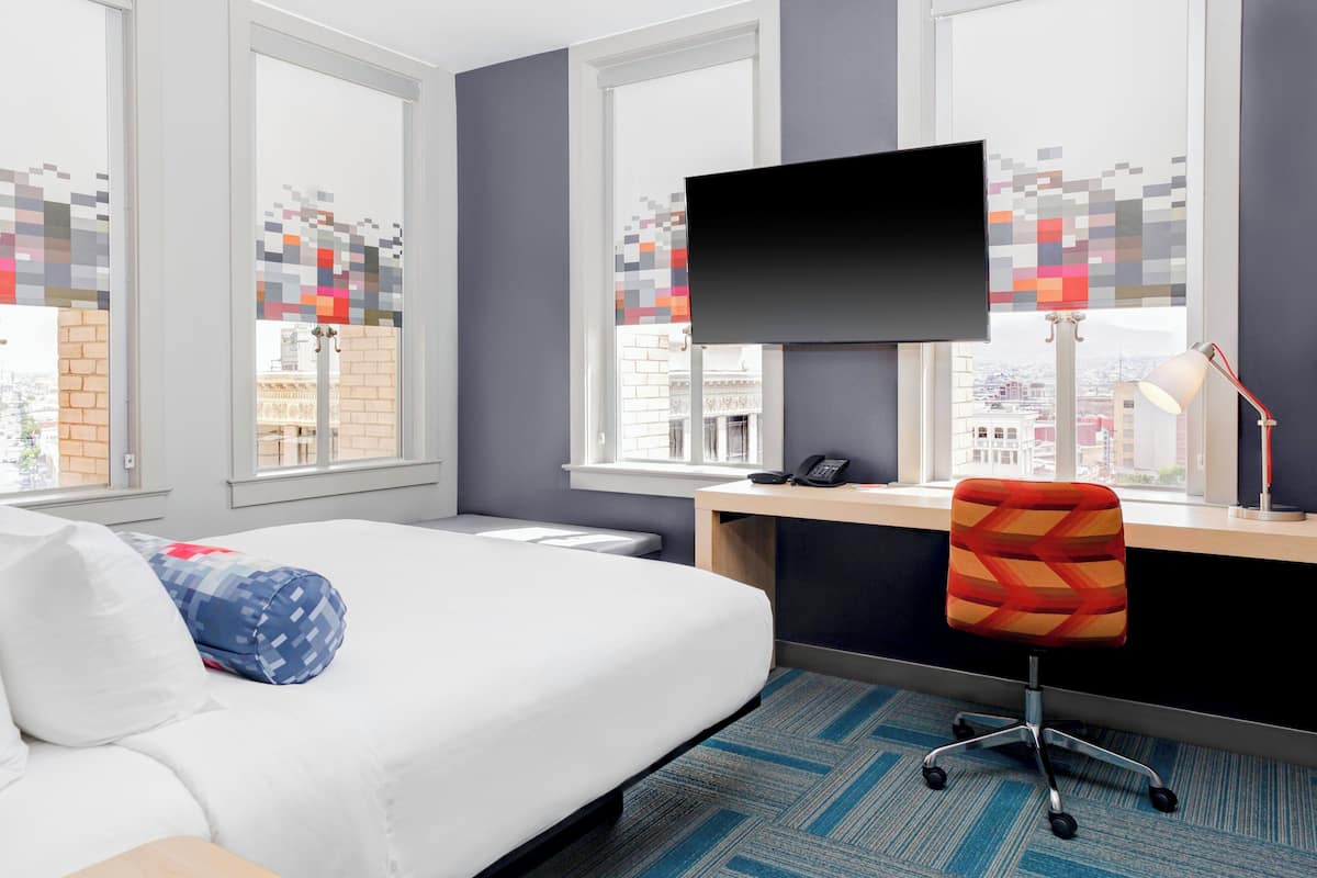 Una habitación de hotel con cama, escritorio con monitor de computadora, silla y ventana con vista a la ciudad.