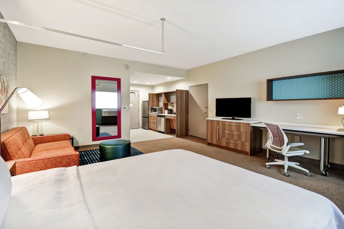 A hotel room with a large bed, a desk with a chair, a TV, a kitchenette, and a bathroom visible through an open door.