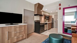 A modern kitchen with a flat-screen TV, built-in oven, microwave, and stainless steel appliances. There is a seating area with a teal ottoman and a patterned rug. - Home2 Suites by Hilton Columbus Downtown (Columbus)
