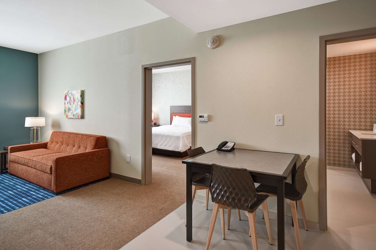 A hotel room with a sofa, a dining table with chairs, a bedside table with a lamp, and a bed with a white comforter.