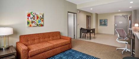 A hotel room with a sofa, a dining table with chairs, a desk with a chair, and a blue patterned rug.