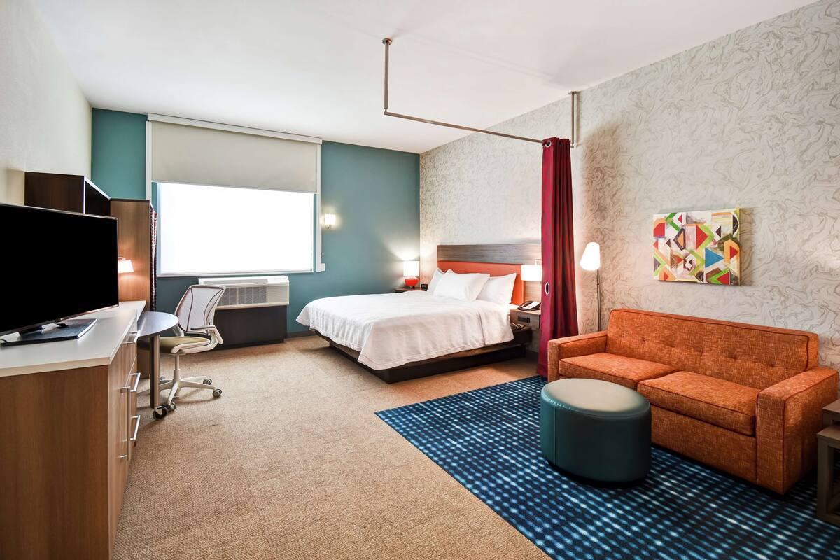A hotel room with a bed, a desk with a computer, a TV, an orange sofa, a patterned rug, and a painting on the wall.