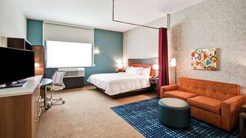 Habitación de hotel con cama, escritorio con computadora, televisor, sofá naranja, alfombra estampada y un cuadro en la pared.