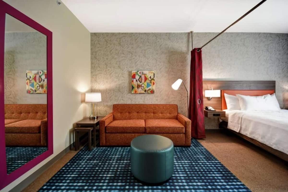 A hotel room with a bed, a sofa, a round ottoman, a mirror, two wall art pieces, and a lamp.
