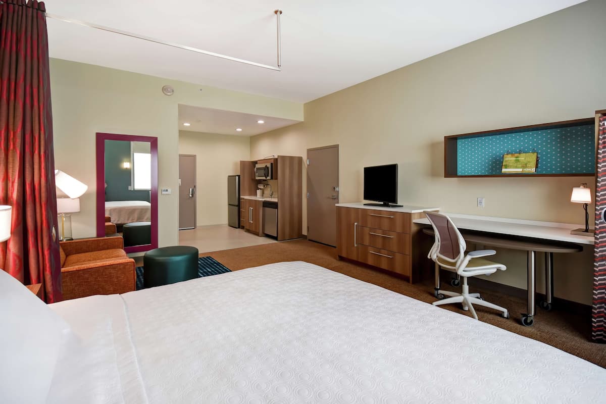 A hotel room with a bed, desk, chair, kitchenette, and a television.