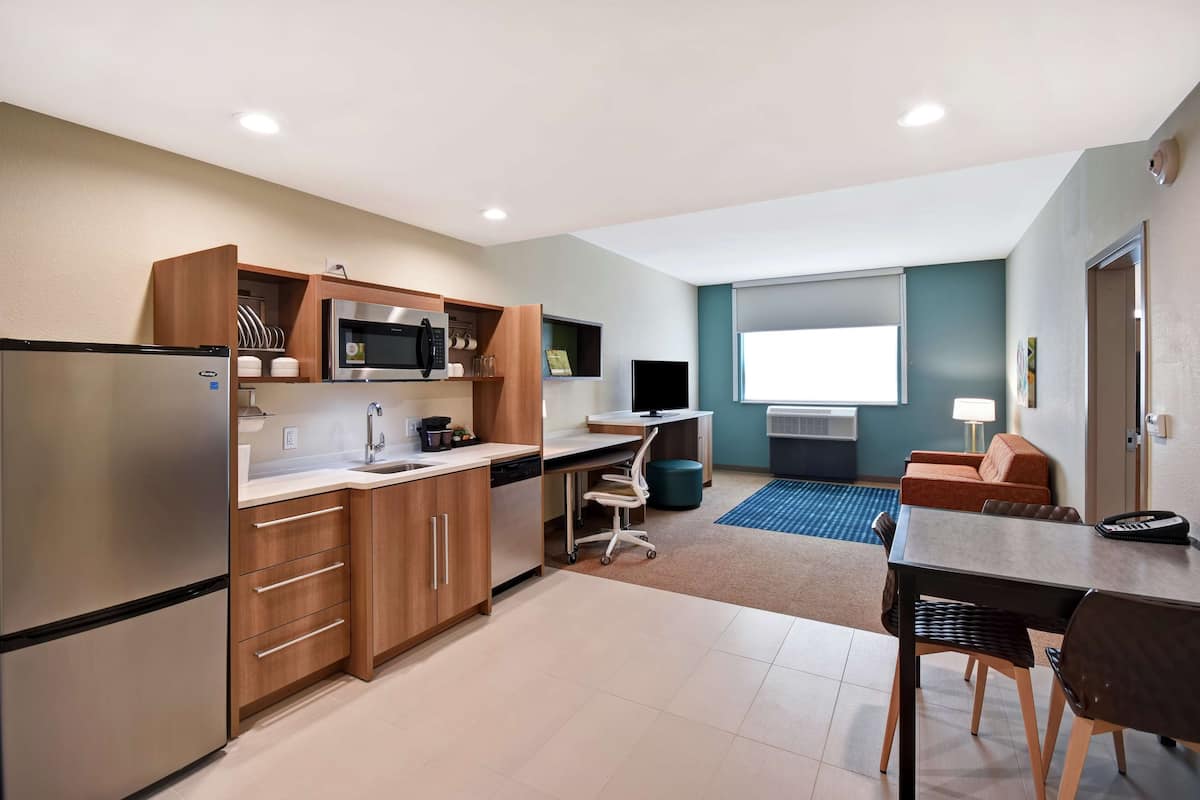A modern kitchen with a refrigerator, microwave, and wooden cabinets. There is a dining area with a table and chairs, and a small seating area with a sofa.